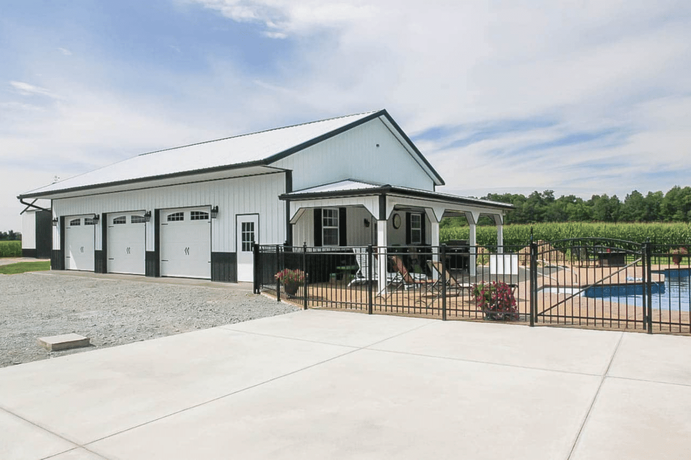 A white 45x100 barndominium kit with black trim, featuring three large garage doors and a covered patio. The patio includes outdoor seating and overlooks a fenced-in pool area with a clear blue swimming pool. The surrounding area has gravel and concrete surfaces, with green fields and trees in the background.