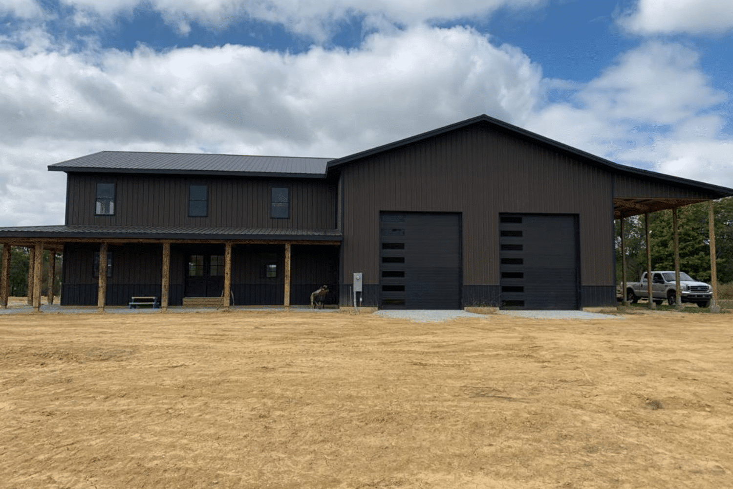 A large 45x100 barndominium kit with a dark brown exterior and black trim, featuring a two-story living area on the left with a covered porch supported by wooden beams. The right side includes two large black garage doors and an extended open-sided structure. The building is surrounded by a dirt lot under a partly cloudy sky with a truck parked to the side.