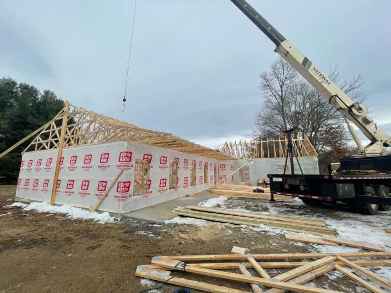 A barndominium under construction with wooden roof trusses being installed. A large crane assists in lifting the trusses onto the structure, which is partially wrapped in protective sheeting. Construction materials and tools are scattered around the snowy ground at the site.