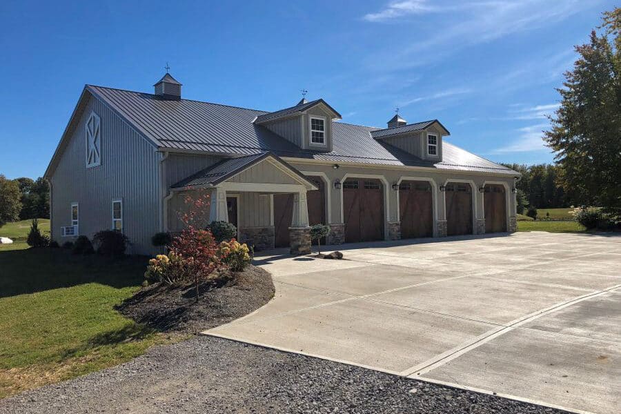 gray pole barn garage with five brown overhead carriage garage doors and two dormers in black metal roof featuring stone detailing along the bottom and a side door with an overhang over it