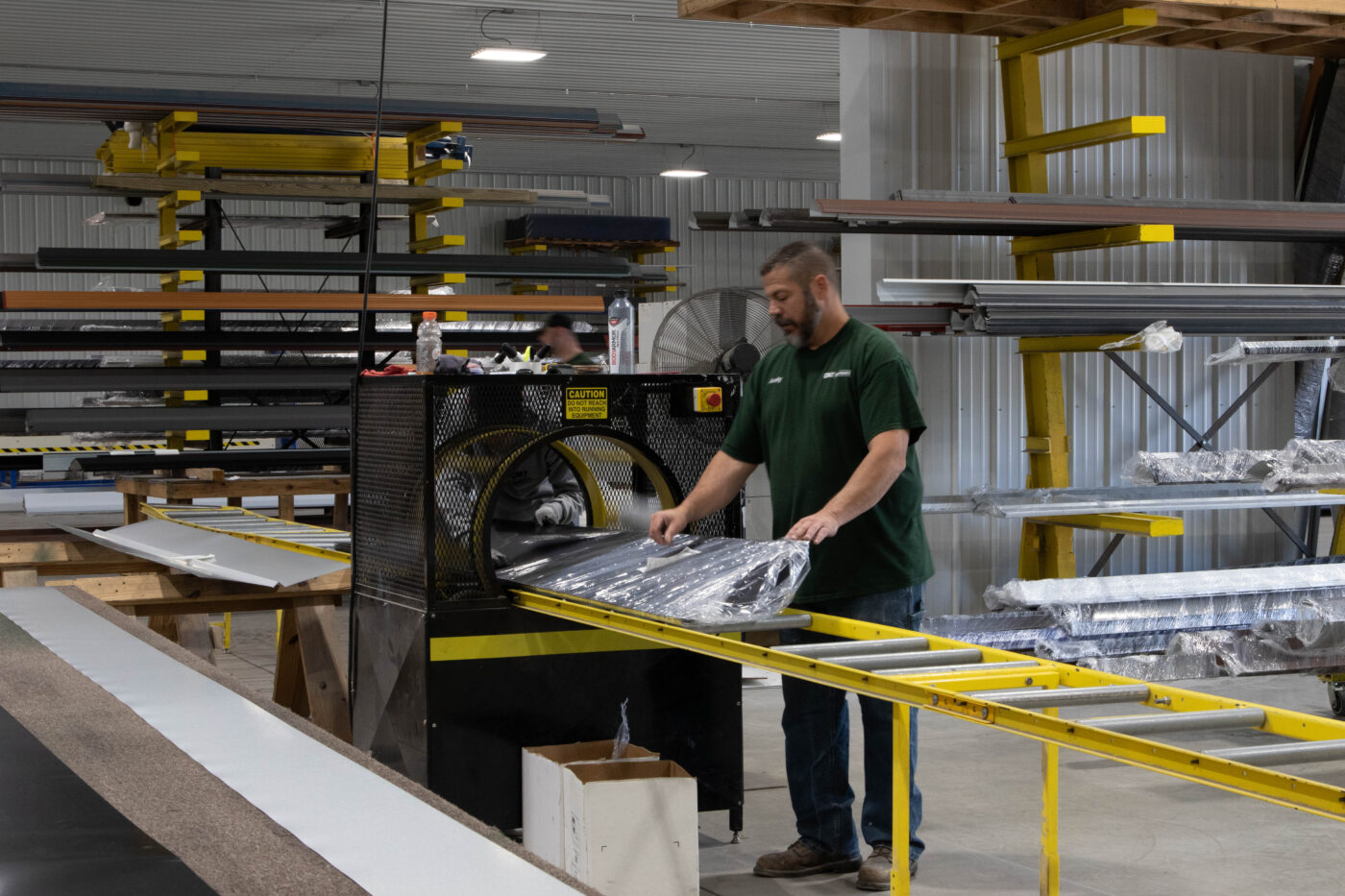 man in green shirt wrapping metal panel in plastic and adding sticker as it comes out of black part of machine