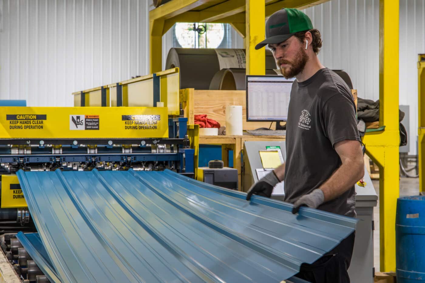 man pulling out blue metal panel from blue and yellpow machine used to custom cut various ribbing into the panel to meet the specification of a 30x48 pole barn garage kit