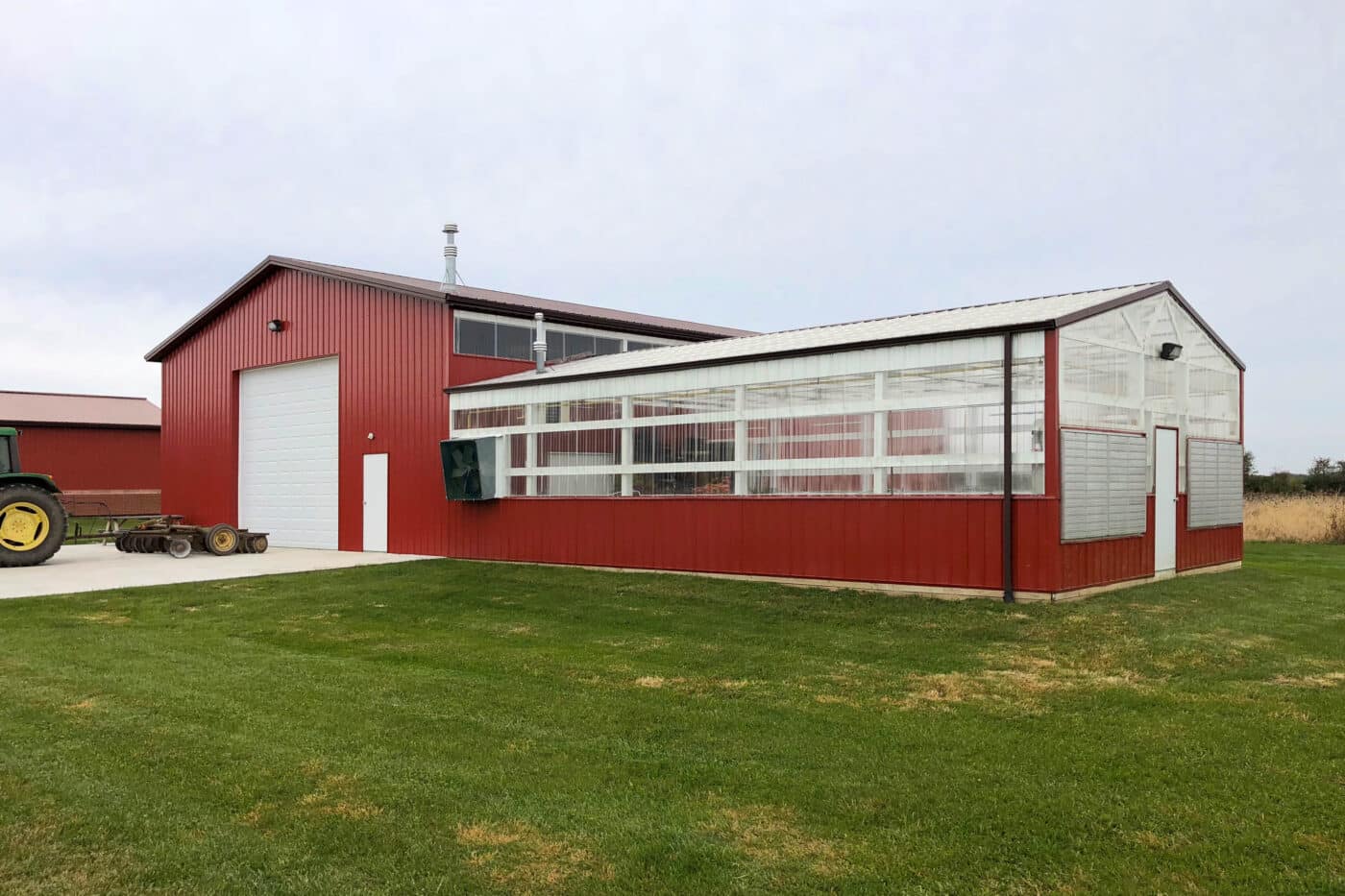 red metal sided 60x80 agricultural pole barn with clera green house siding as well featuring one white overhead door with tractor parked in front