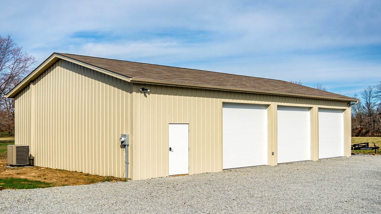 light yellow 30x48 pole barn garage with brown shingle roof three white overhead doors and a white side door along gravel drive